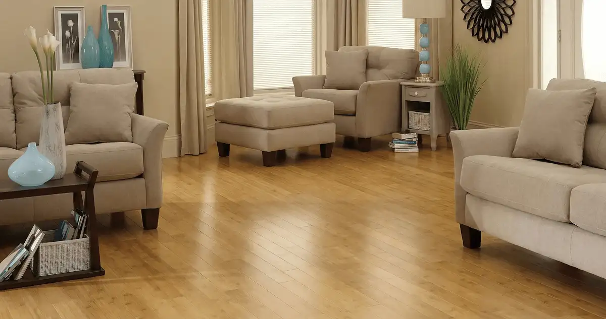 A living room with professionally installed bamboo flooring, beige sofas, a matching ottoman, a side table with a blue lamp, vases, framed pictures, and large windows with blinds and curtains in Yonkers, NY